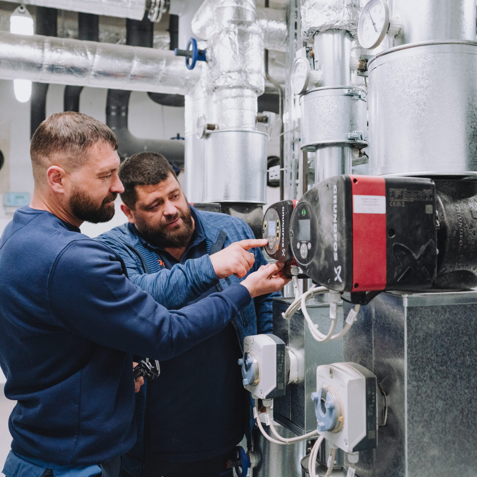 Zwei Männer in Arbeitskleidung betrachten eine Anzeige an einer Heizungsanlage