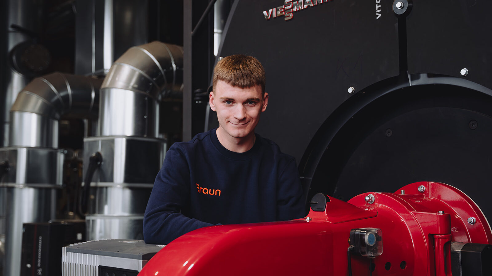 Ein junger Mann in Braun-Arbeitskleidung vor einem modernen Heizkessel