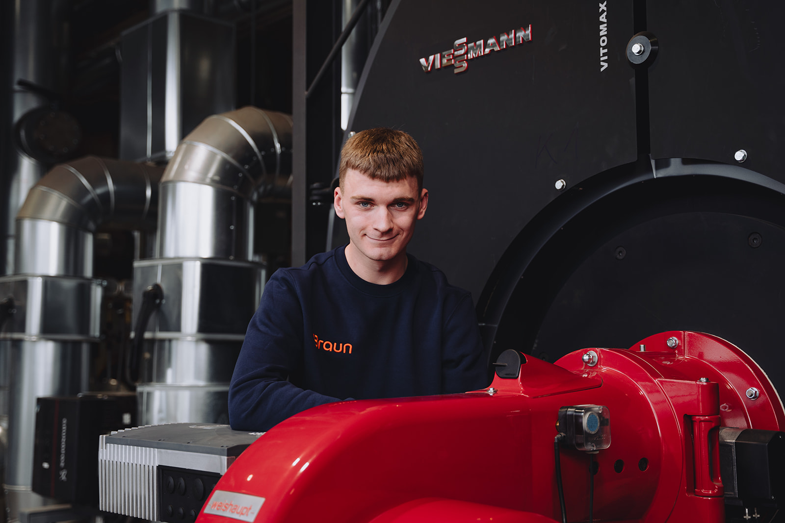 Ein junger Mann in Braun-Arbeitskleidung vor einem modernen Heizkessel