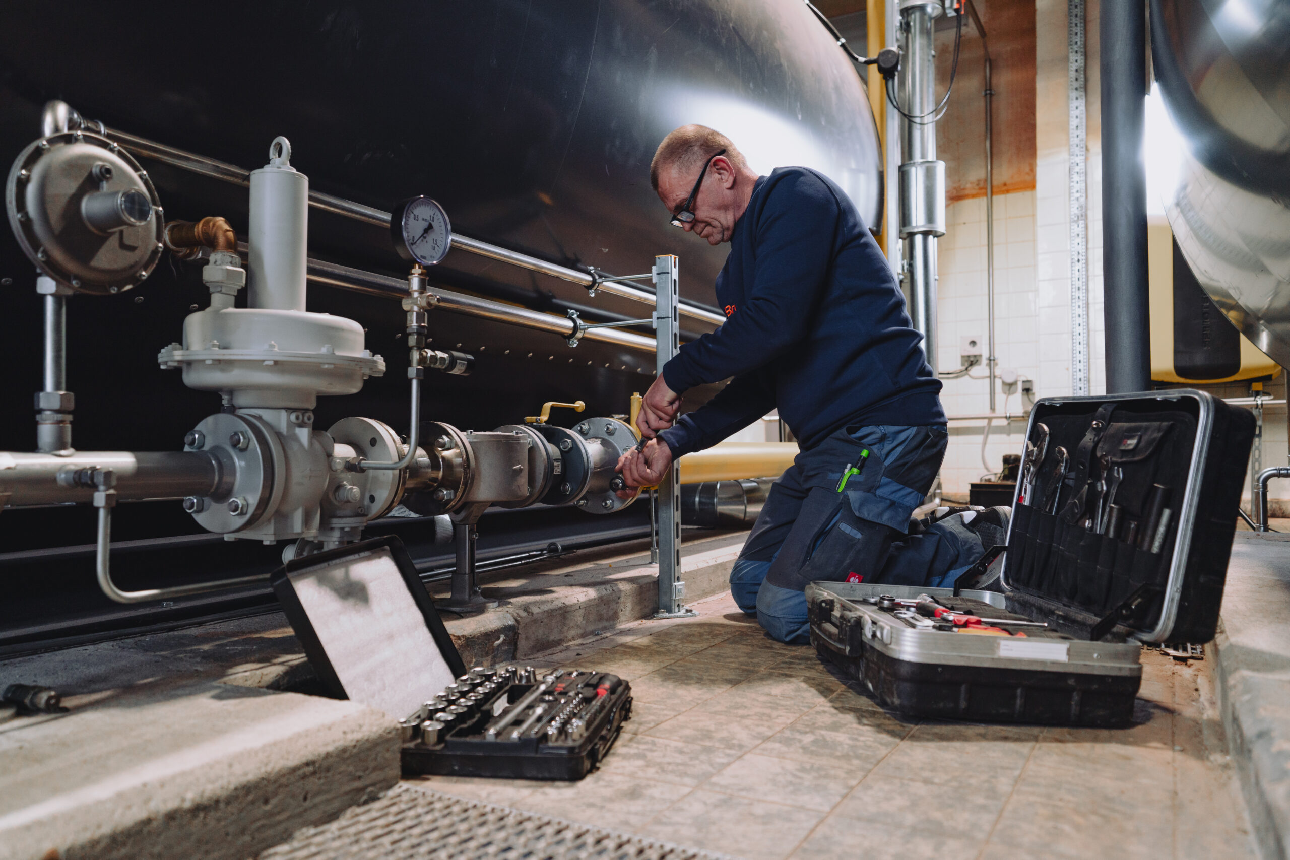 Ein Mitarbeiter von Braun in Arbeitskleidung zieht auf dem Boden kniened eine Mutter an einer Rohrleitung fest.